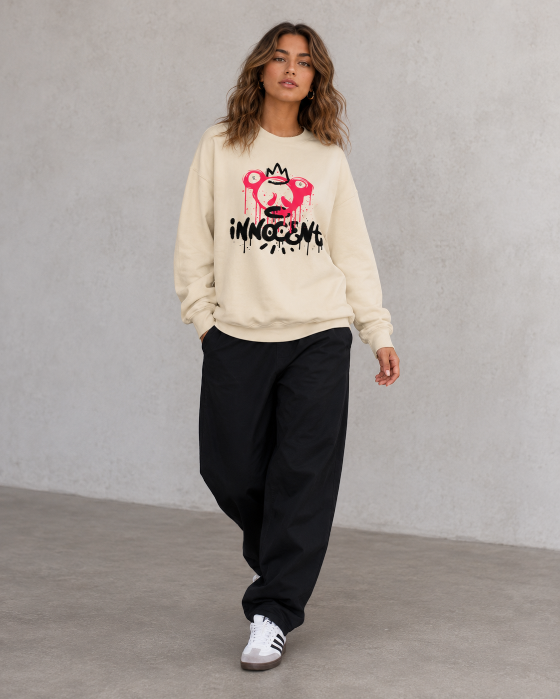 Person wearing a beige sweatshirt with a graphic design and text, black pants, and white sneakers on a plain background