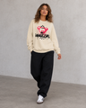 Person wearing a beige sweatshirt with a graphic design and text, black pants, and white sneakers on a plain background