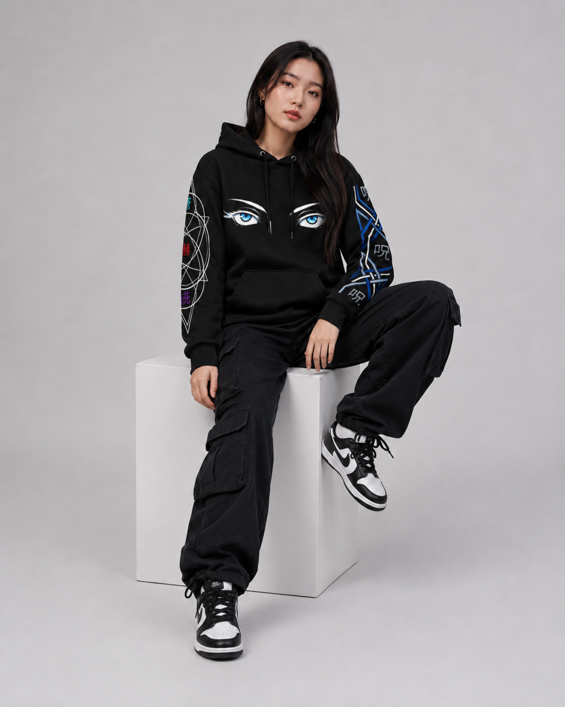 Person wearing a black hoodie with eye designs, sitting on a white cube against a gray background