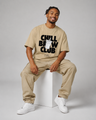Man sitting on a white stool wearing a beige 'Chill Brew Club' t-shirt and pants with white sneakers.