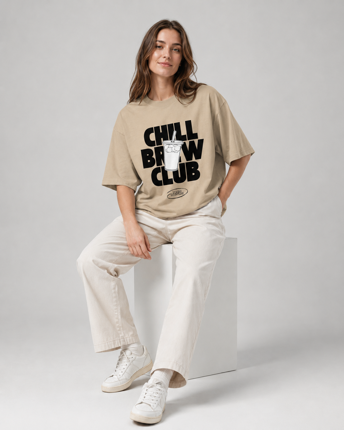 Woman wearing a beige t-shirt with 'Chill Brew Club' text, sitting on a white block against a gray background
