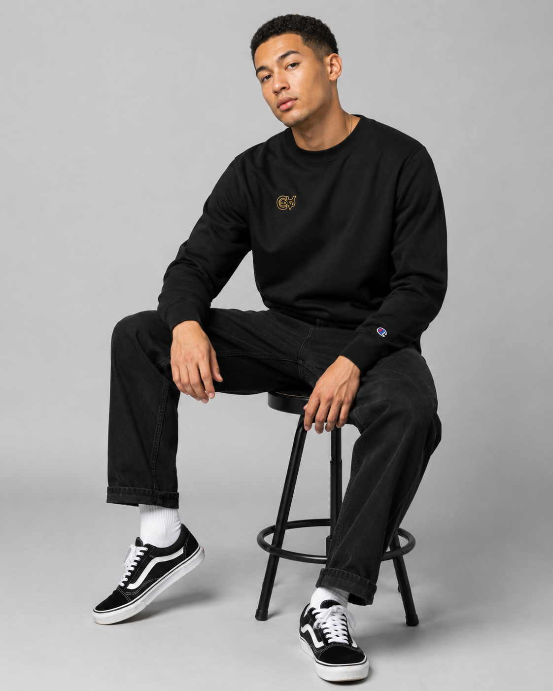 Person wearing a black sweatshirt and pants sitting on a stool against a gray background