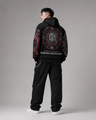 Person wearing a black hoodie with intricate designs on the back, standing against a gray background