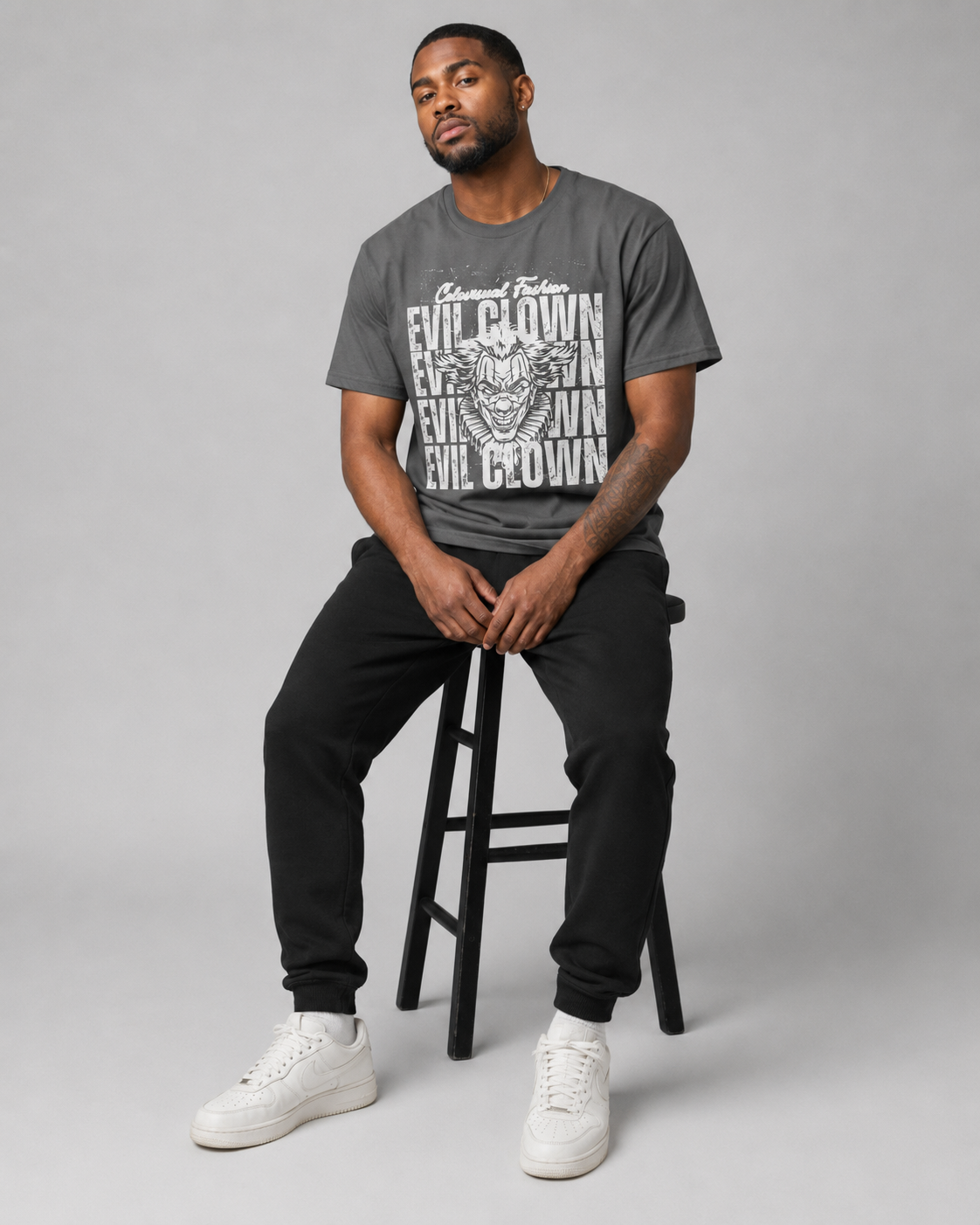 Man sitting on a stool wearing a gray t-shirt with text and black pants on a plain background