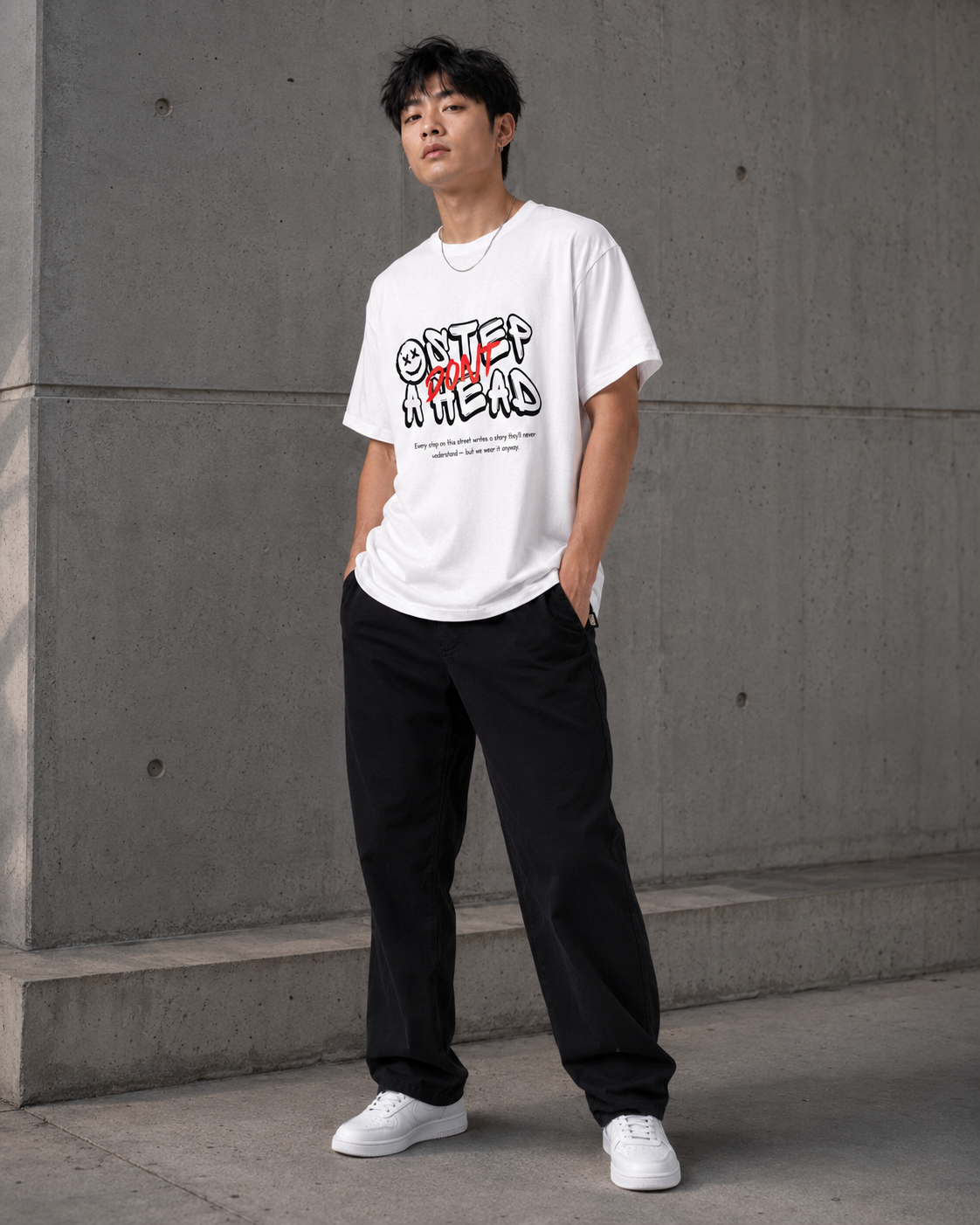 Person wearing a white t-shirt with text and black pants standing against a gray wall.