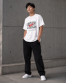 Person wearing a white t-shirt with text and black pants standing against a gray wall.