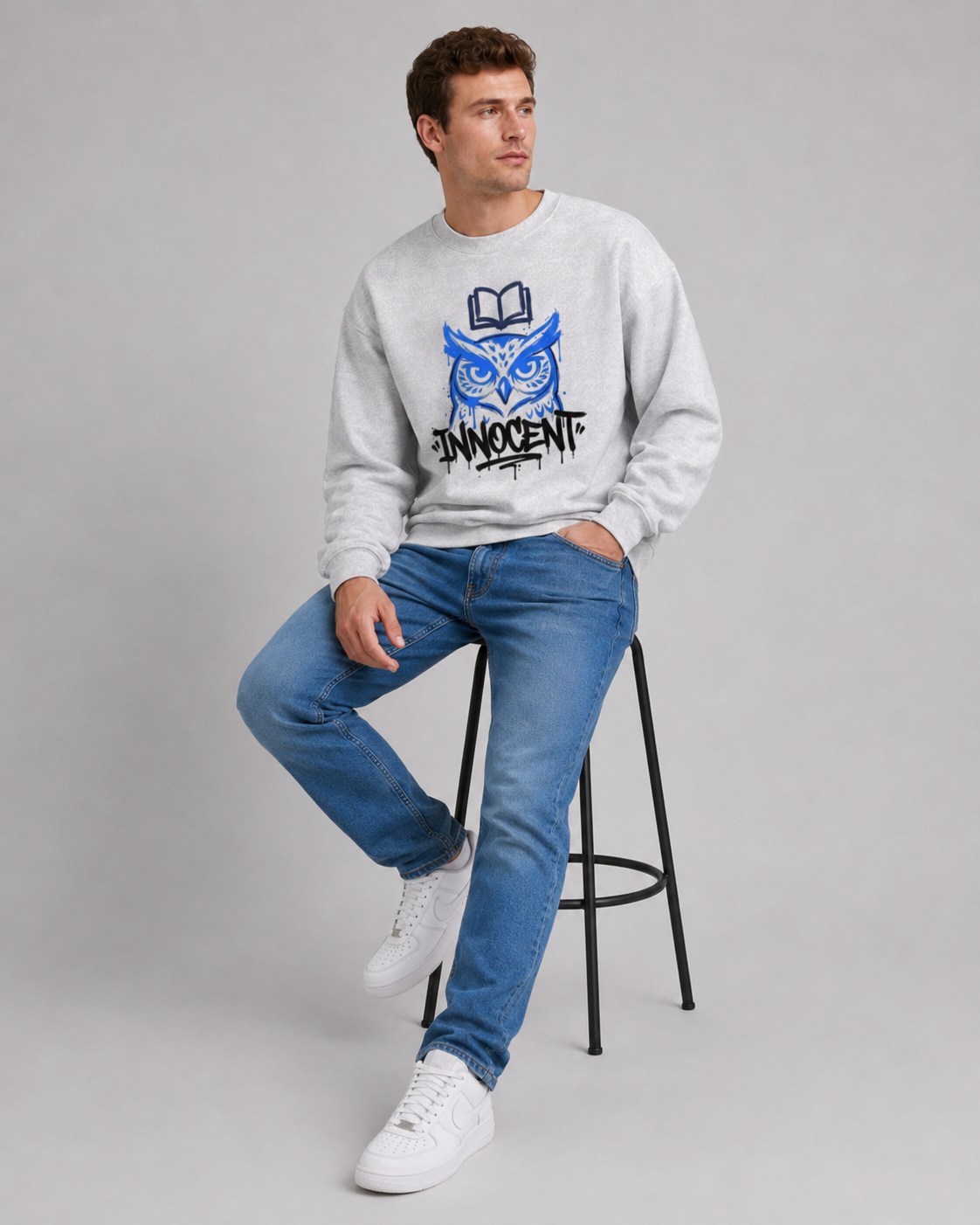 Man wearing a gray sweatshirt with a blue owl graphic and text, sitting on a stool against a gray background.