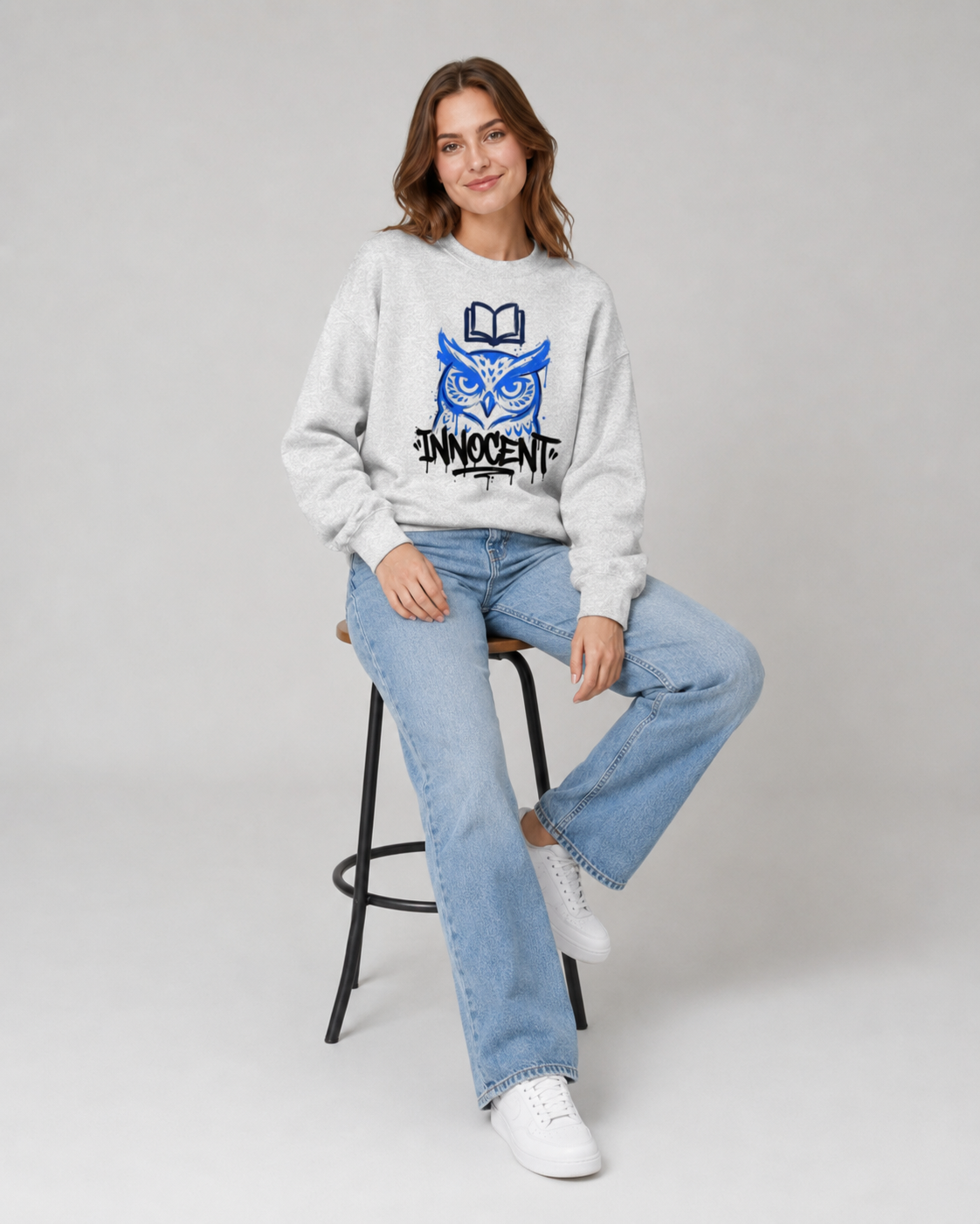 Woman wearing a white sweatshirt with a blue graphic design, sitting on a black stool against a gray background.