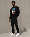 Man wearing a black sweatshirt with a colorful graphic design, black cargo pants, and white sneakers against a concrete wall.