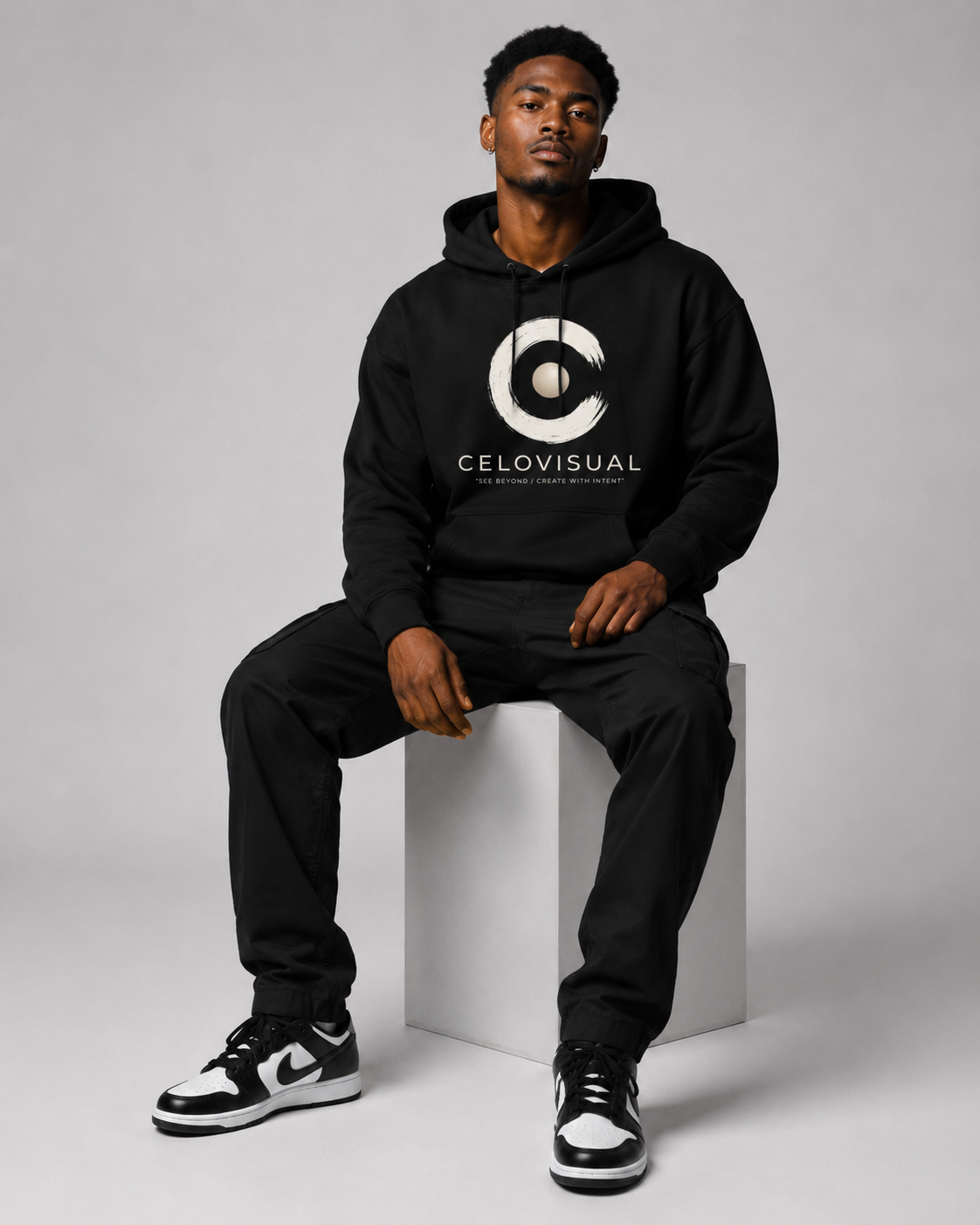 Man wearing a black hoodie with a logo and text, sitting on a white cube against a gray background.