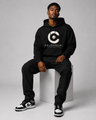 Man wearing a black hoodie with a logo and text, sitting on a white cube against a gray background.