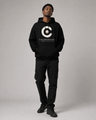 Person wearing a black hoodie with a logo and text, standing against a plain background