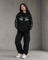 Person wearing a black hoodie with eye design and matching pants against a concrete wall.