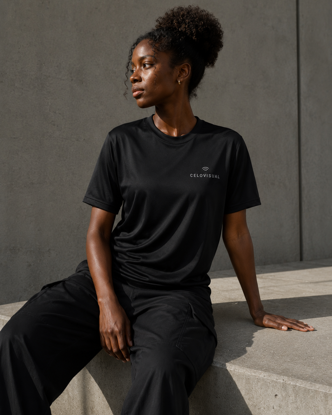 Person wearing a black t-shirt with a brand logo, sitting against a gray wall.