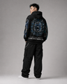 Person wearing a black hoodie with intricate designs and text, standing against a gray background
